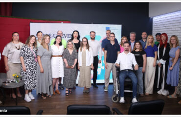 Grup de persoane la un eveniment corporativ, stând în fața unui panou de prezentare într-o cameră cu pereți roșii.